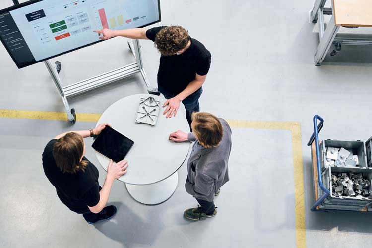 Three employees stand in production and discuss the key figures on a large monitor in store floor management
